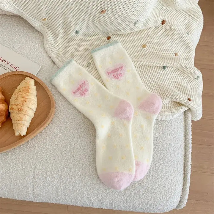 Kósýsokkar - hvítir og bleikir með röndum og teiknimyndahöfði á teppi við hliðina á croissant á borði.